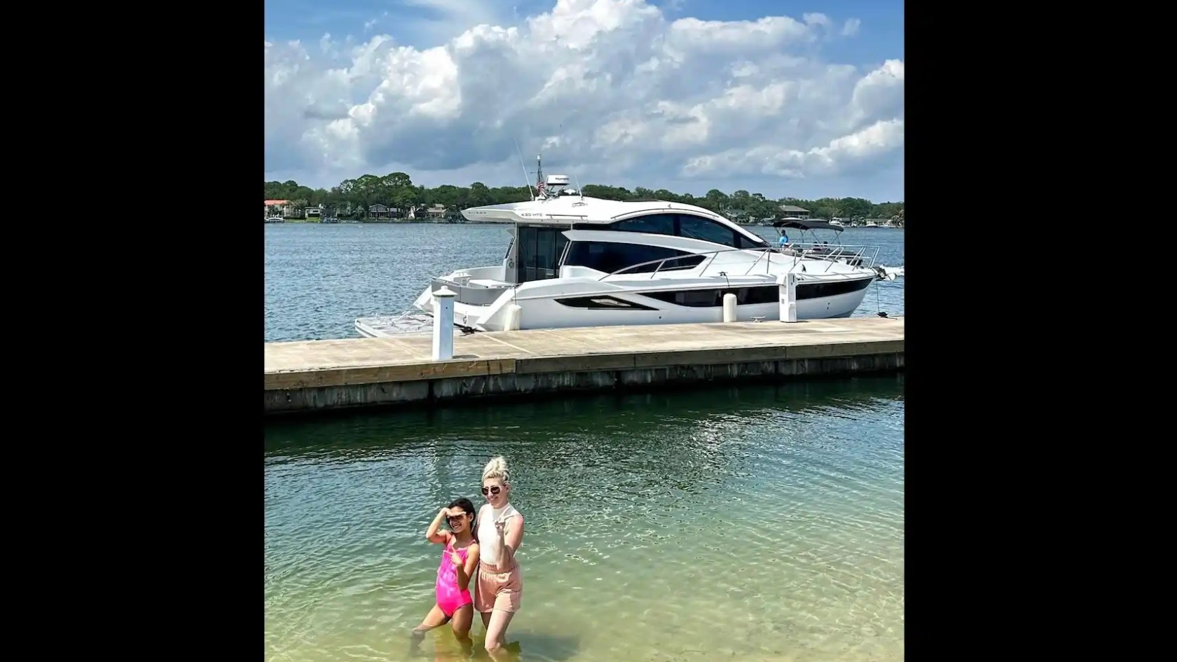 Two people posing in shallow water near a docked yacht under a cloudy sky.