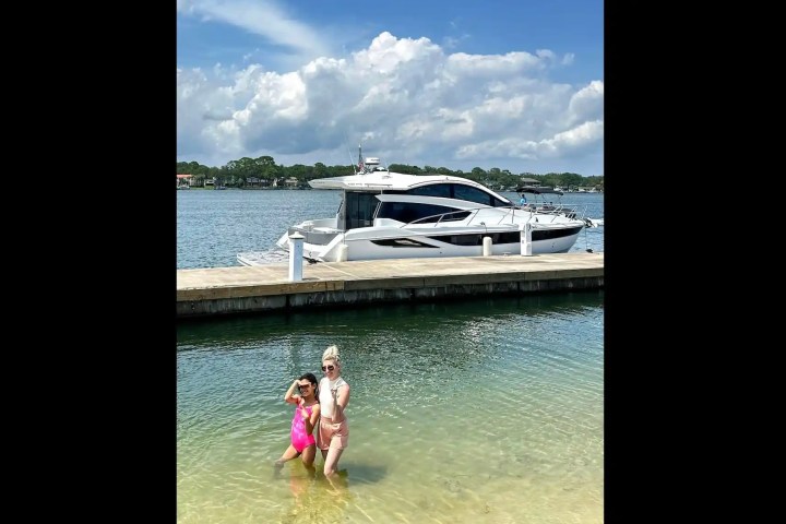 Two people posing in shallow water near a docked yacht under a cloudy sky.