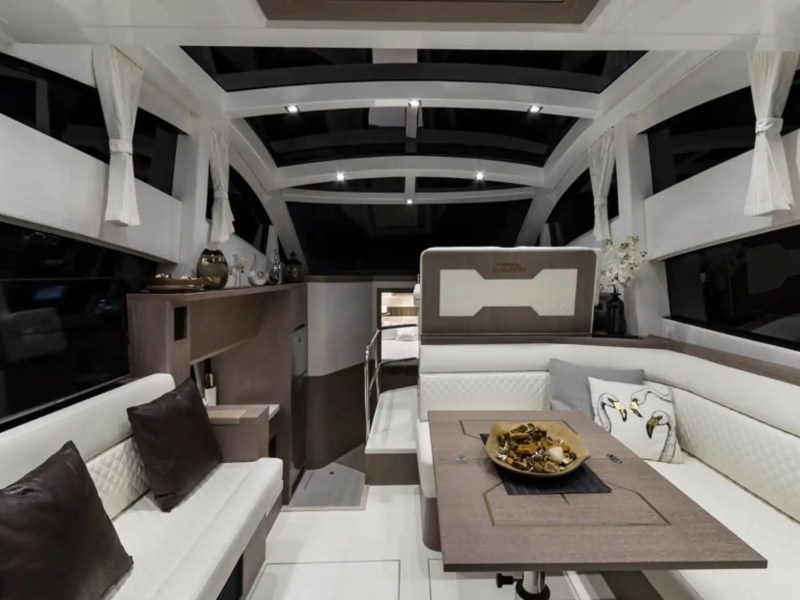 Modern yacht interior with white sofas, brown table, decorative pillows, and large windows.