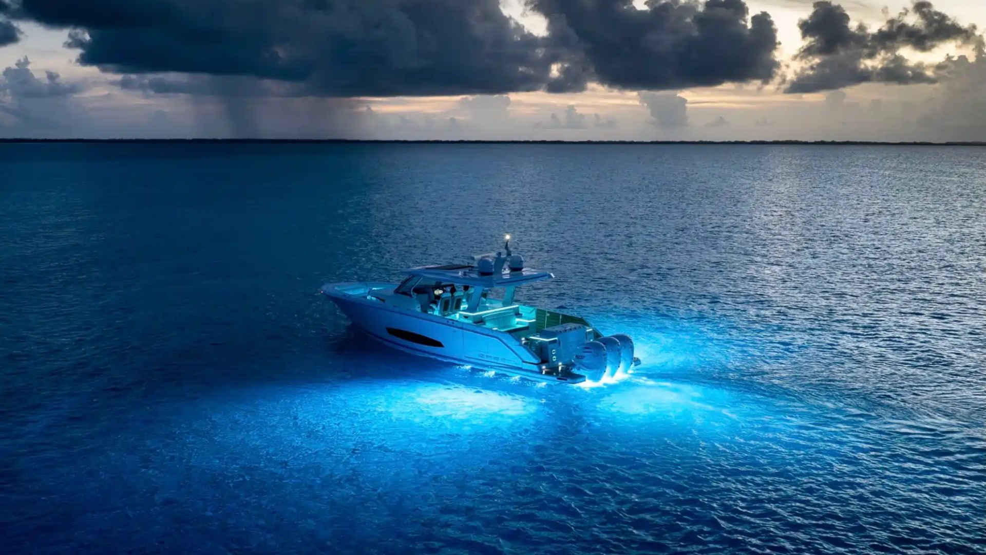 Boat with blue underwater lights on a calm ocean under a dramatic cloudy sky.
