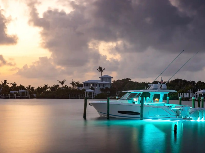 Boat with glowing underwater lights docked at sunset with cloudy sky.