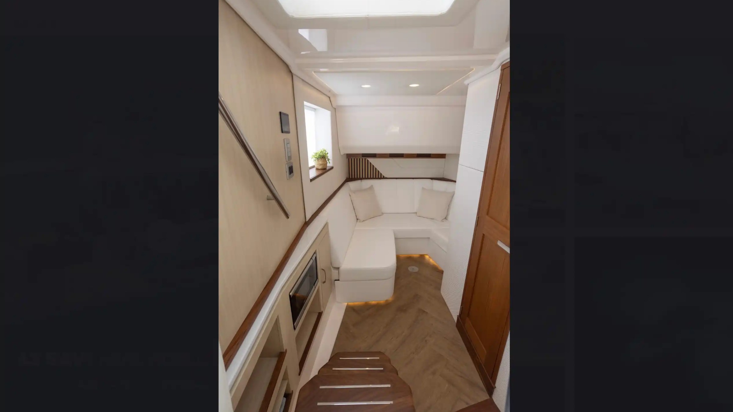 Modern boat cabin with L-shaped beige sofa, wooden stairs, and overhead lighting.