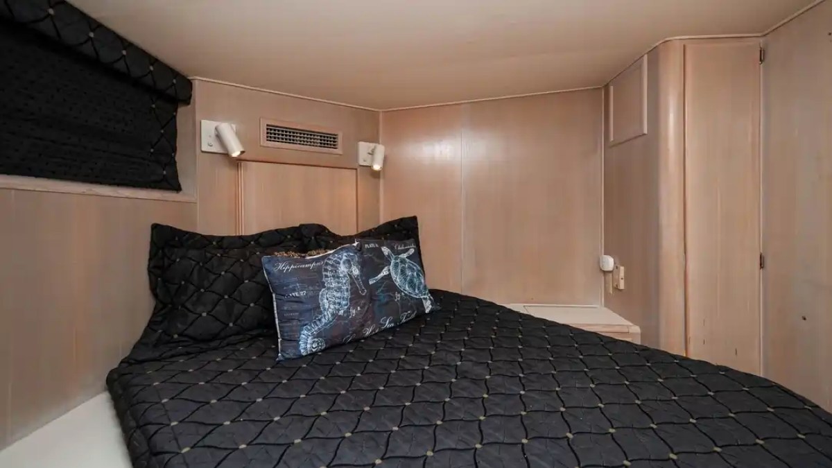Small cozy bed with black quilted bedding and seahorse pillow in a compact, wood-paneled room.