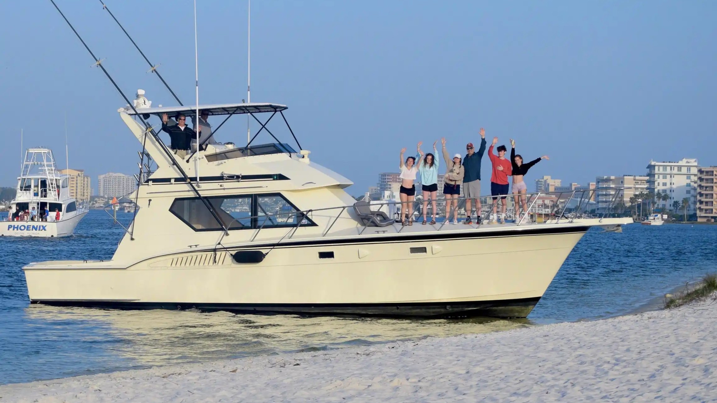 A group of people waving on a yacht near a sandy shore with buildings in the background.