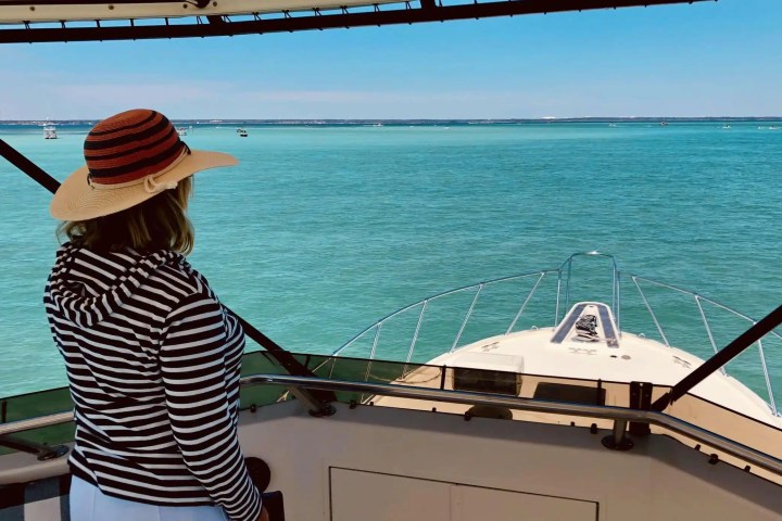 Woman in striped shirt and hat on yacht, looking at turquoise sea and distant boats.