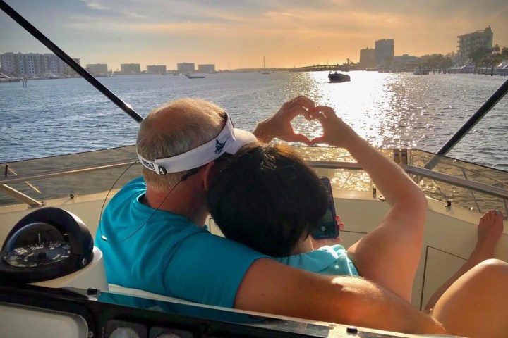 Couple on boat at sunset, making heart shape with hands, overlooking water.