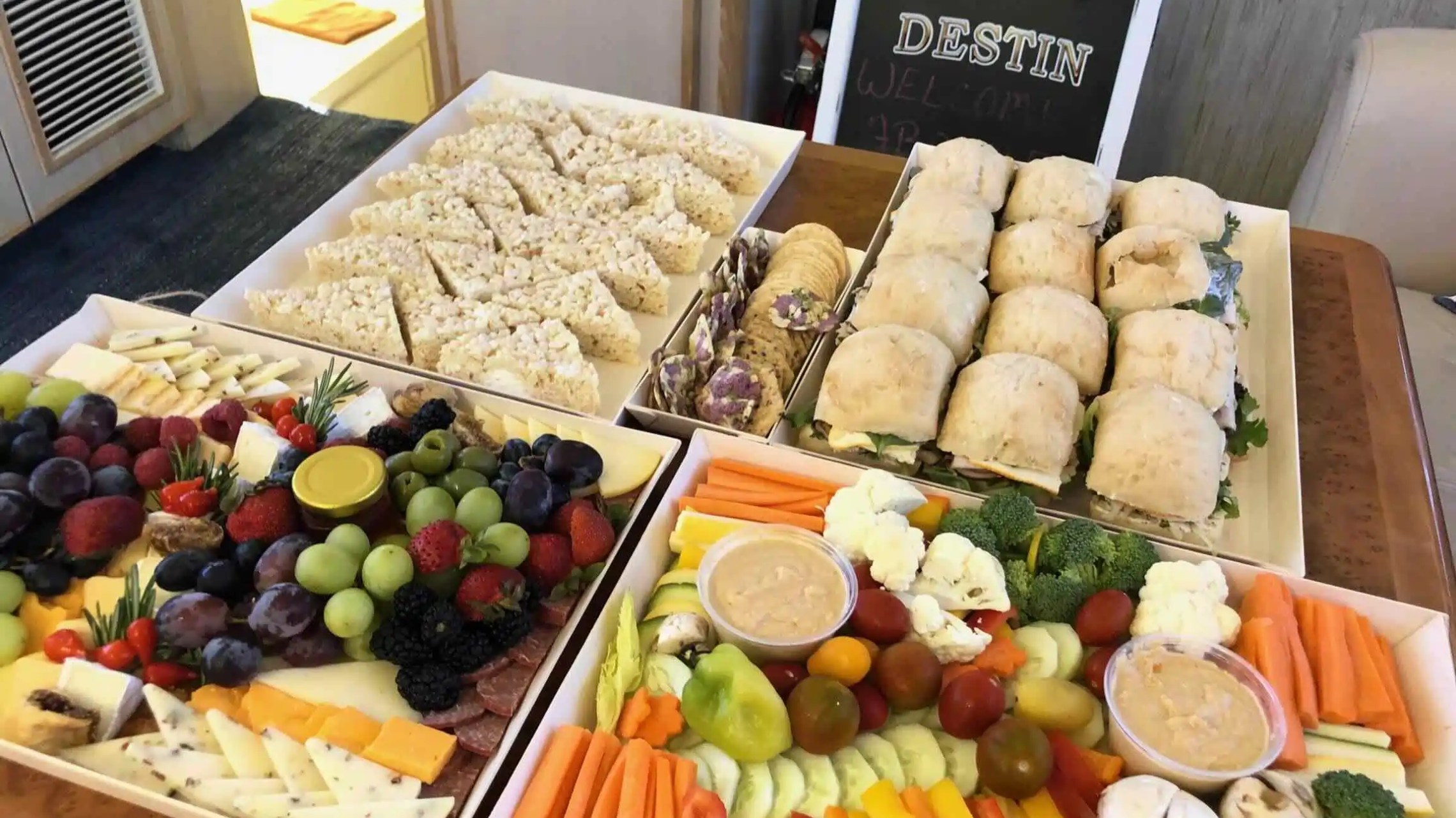 Assorted platters of fruits, vegetables, sandwiches, and rice treats with a welcome sign in background.