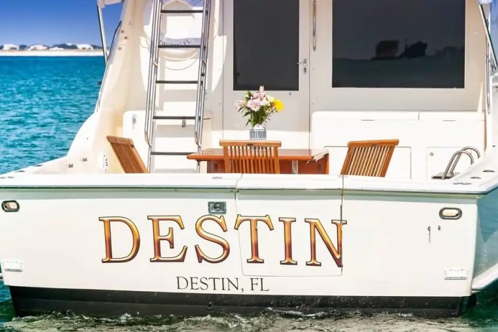 The back of a boat named 'Destin' with a table and chairs on deck, floating in blue water.