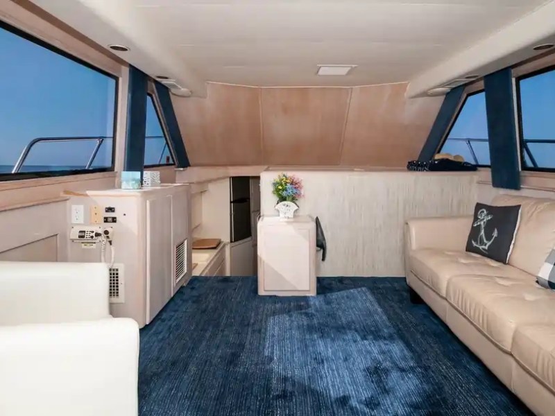 Boat cabin interior with beige couch, armchair, blue carpet, and large windows overlooking the sea.