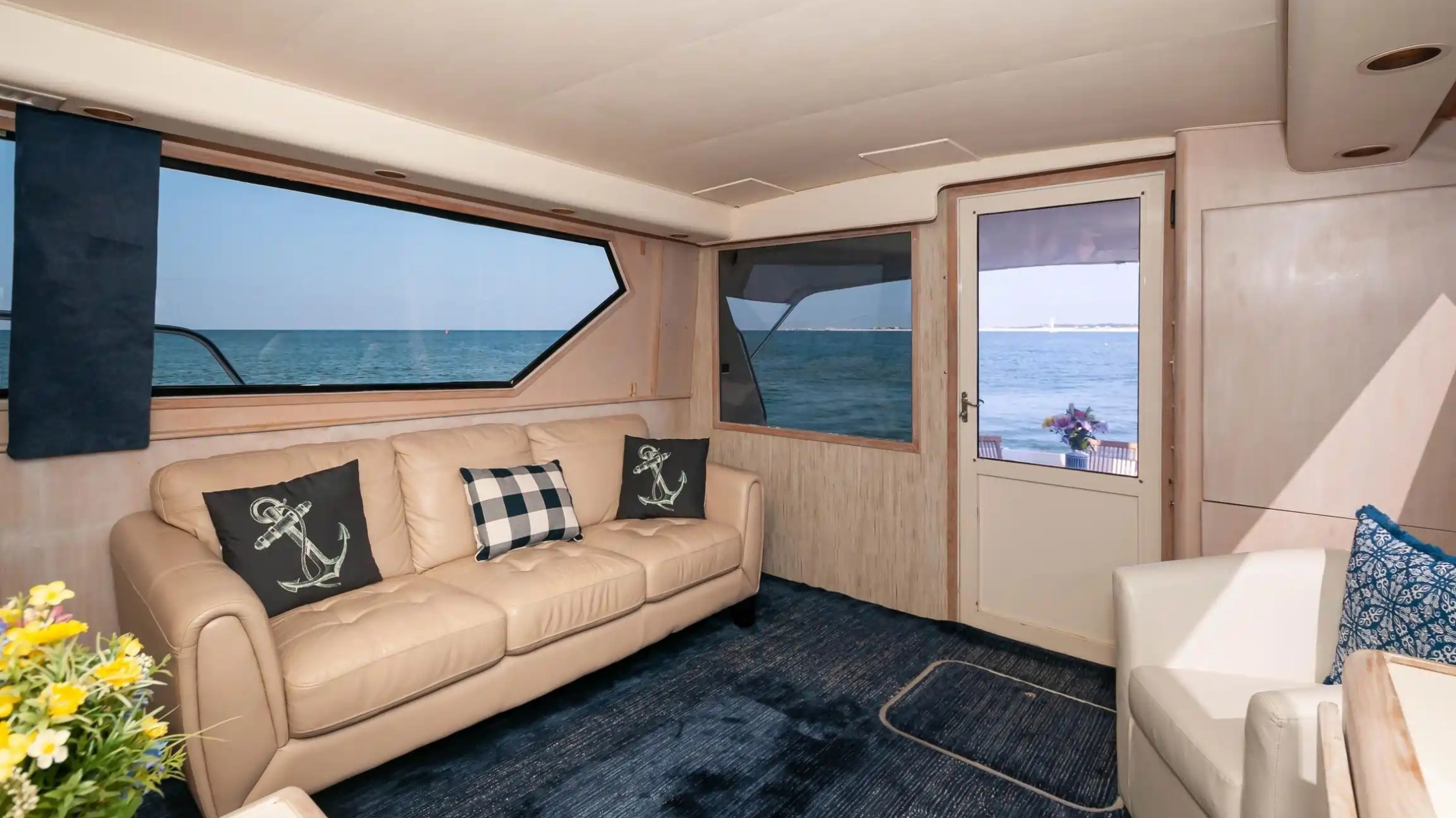 Yacht interior with beige sofa, anchor pillows, blue rug, and ocean view.
