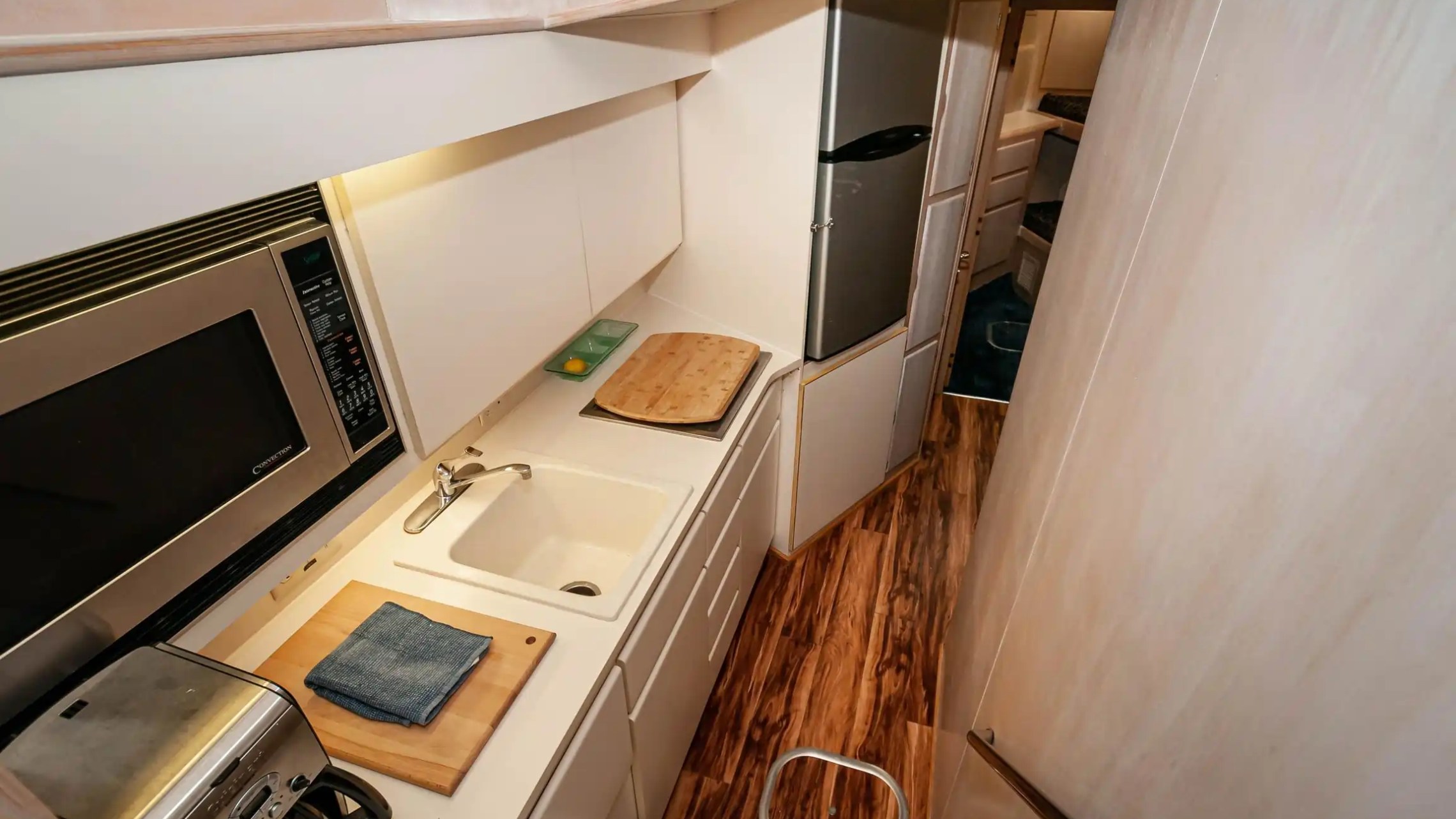 Narrow kitchen with microwave, sink, toaster, cutting board, and fridge, on a wooden floor.