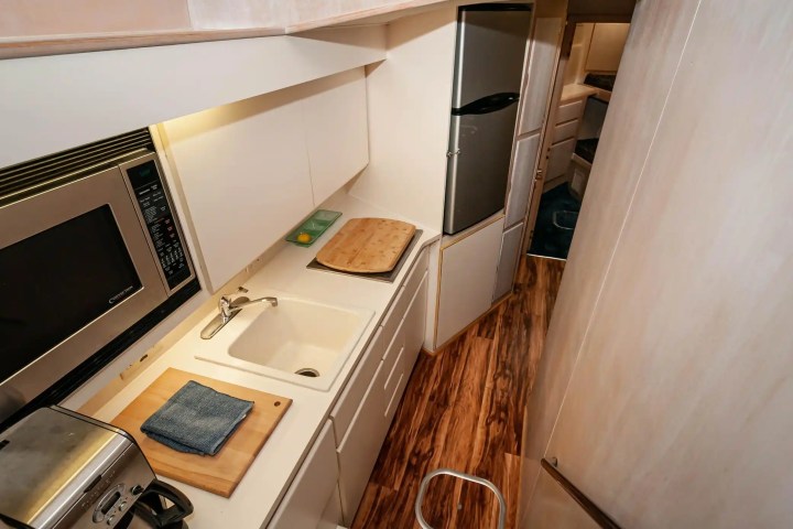 Narrow kitchen with microwave, sink, toaster, cutting board, and fridge, on a wooden floor.