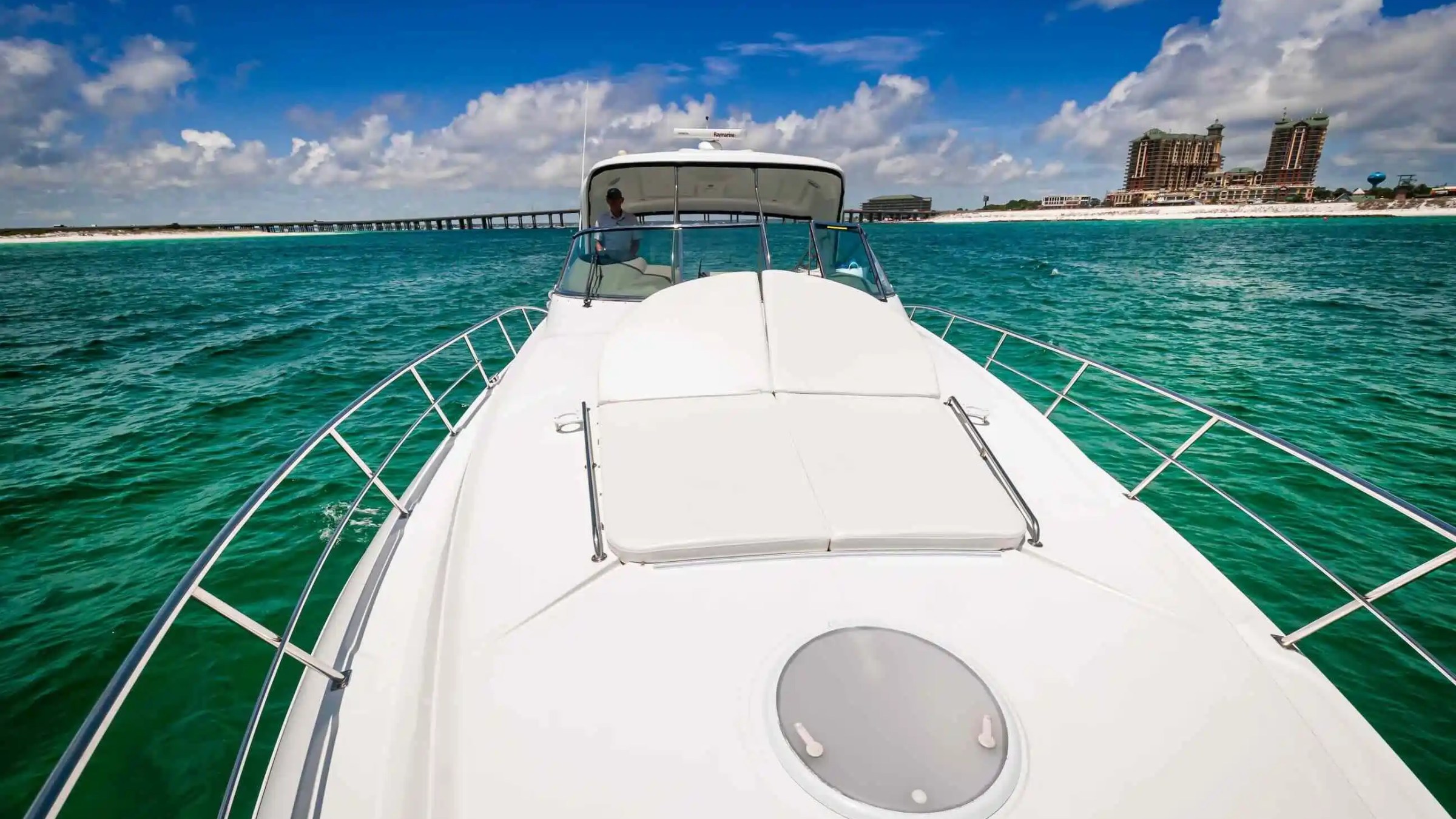 View from yacht deck toward beach with buildings and pier in the distance on a sunny day.