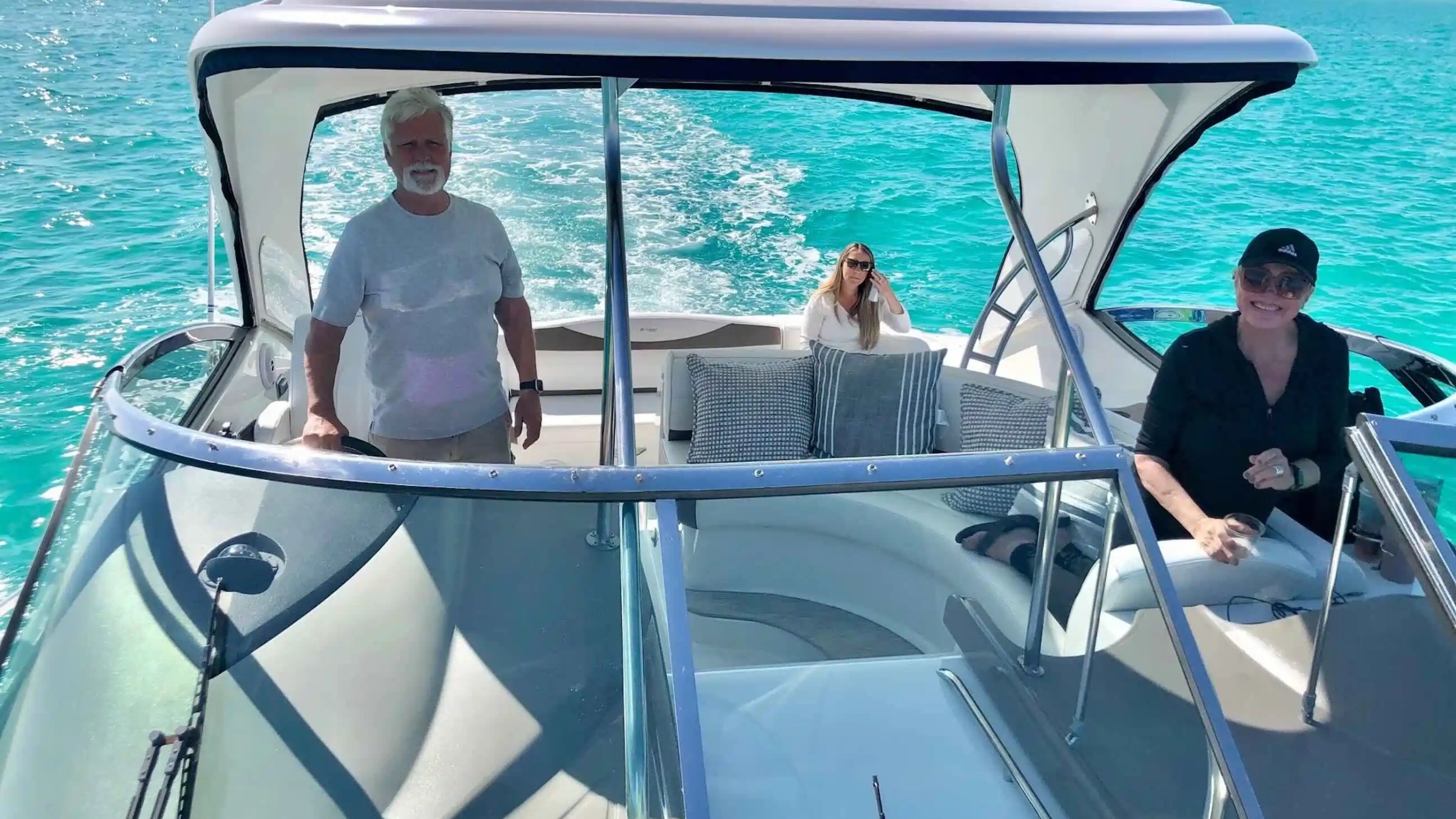 Three people on a motorboat in clear blue water, one driving and two sitting, smiling and enjoying the ride.