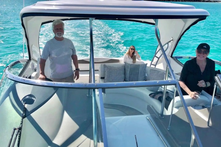 Three people on a motorboat in clear blue water, one driving and two sitting, smiling and enjoying the ride.