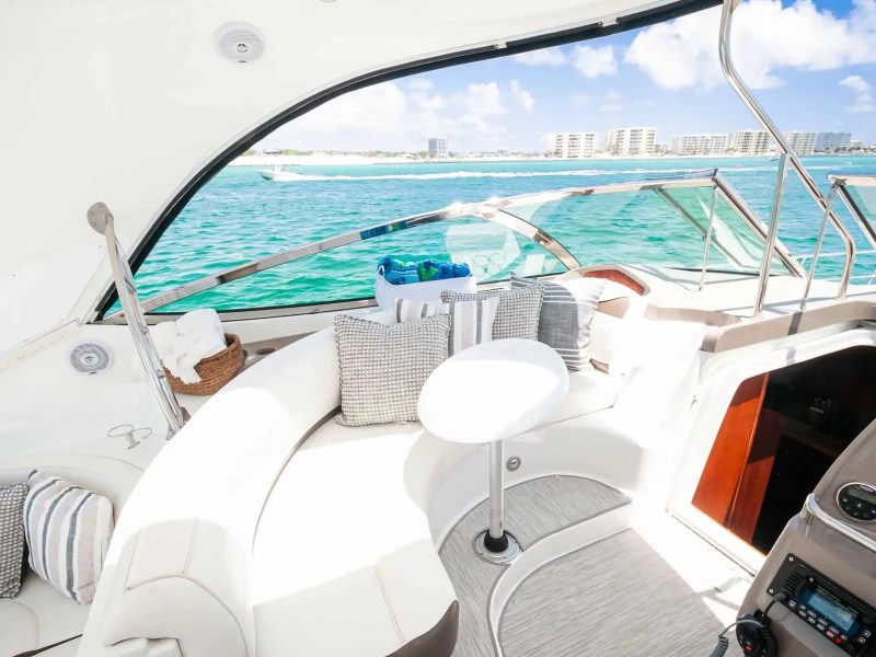 Interior of a yacht with white seating and striped pillows, overlooking blue water and a city skyline.