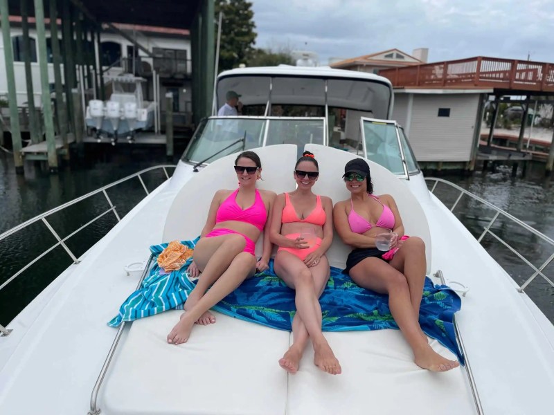 Three people in swimsuits relax on a boat deck with towels on a cloudy day.