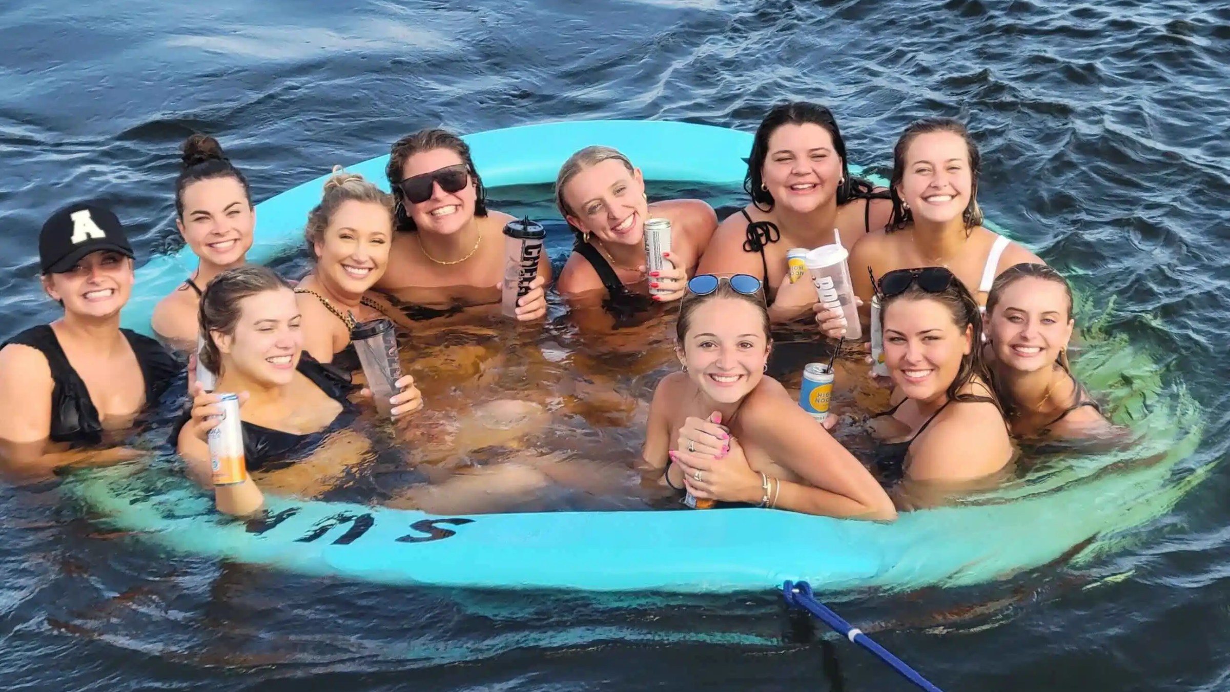 Group of smiling women in swimsuits sitting in a floating ring on the water, each holding a drink.