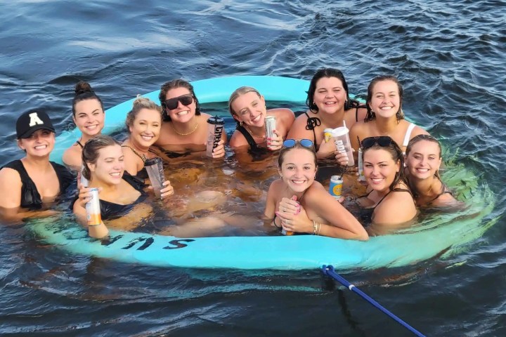 Group of smiling women in swimsuits sitting in a floating ring on the water, each holding a drink.