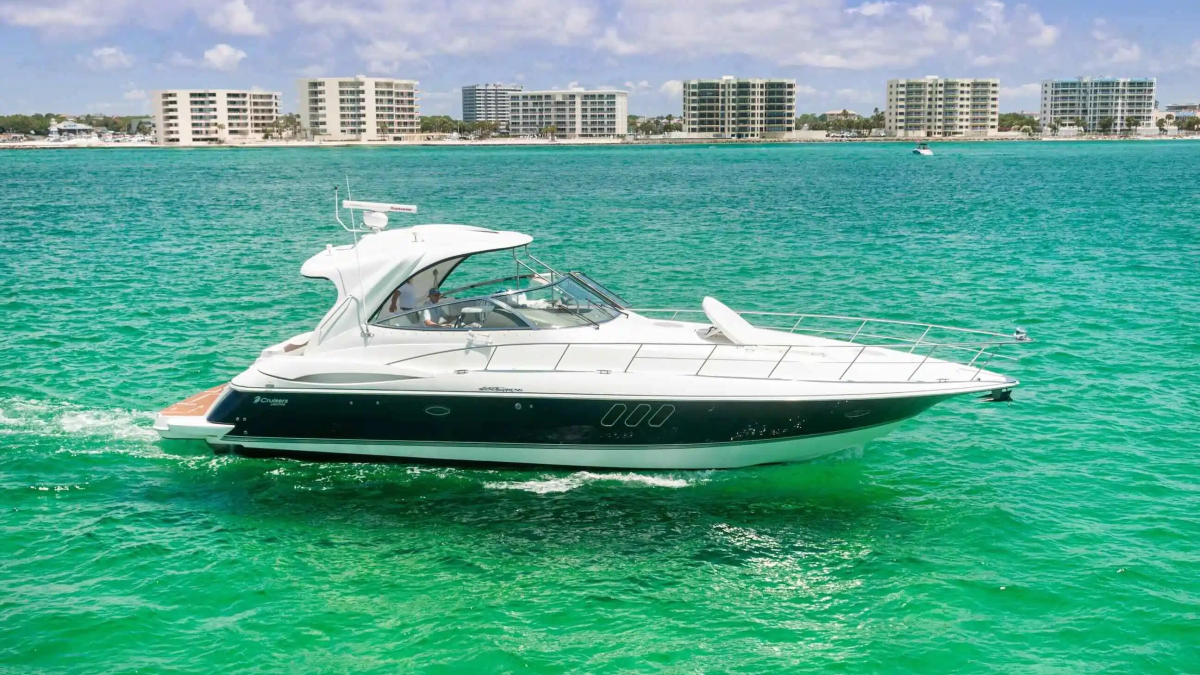 Motor yacht cruising on turquoise water near high-rise buildings on a sunny day.