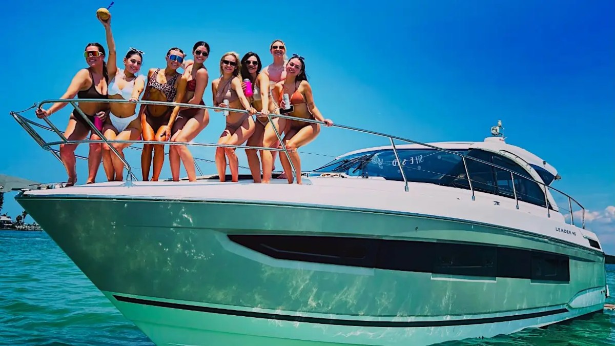 Group of women in swimsuits on yacht holding drinks under clear blue sky.