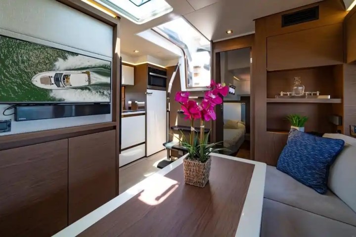 Modern yacht interior with dining table, TV, and pink flowers, featuring wood accents and natural light.
