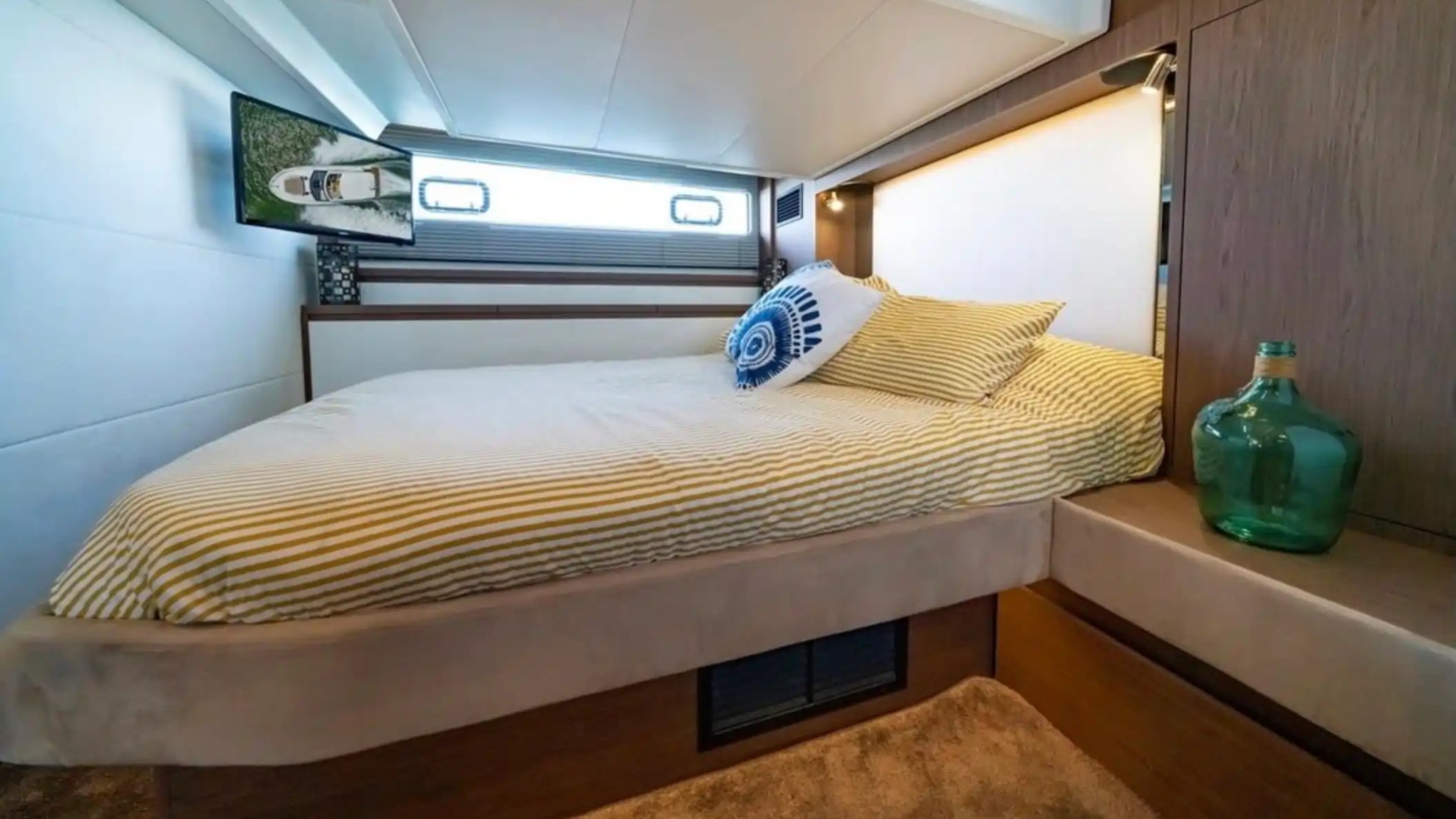 Yacht bedroom with striped bedding, decorative pillow, TV, and a green glass bottle on a wooden surface.
