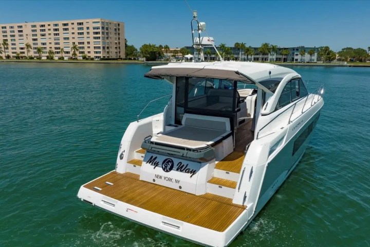 White yacht named 'My Way' in calm water near a row of palm trees and buildings.