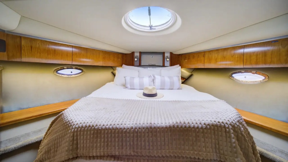 Compact boat cabin with bed, portholes, and skylight; hat on bed, wooden cabinets.