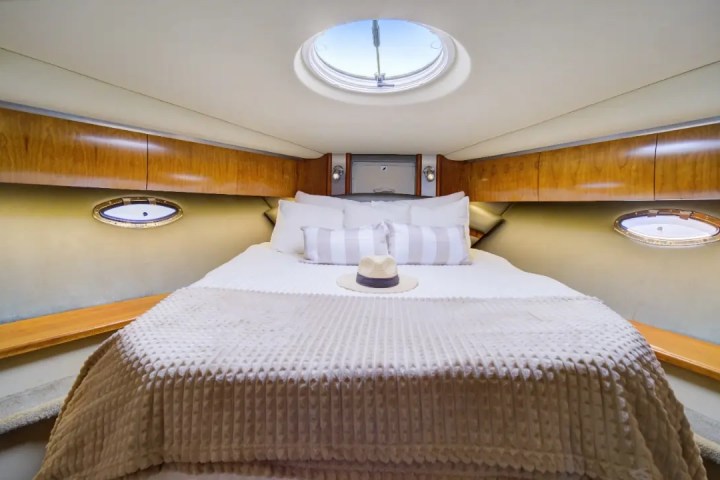 Compact boat cabin with bed, portholes, and skylight; hat on bed, wooden cabinets.