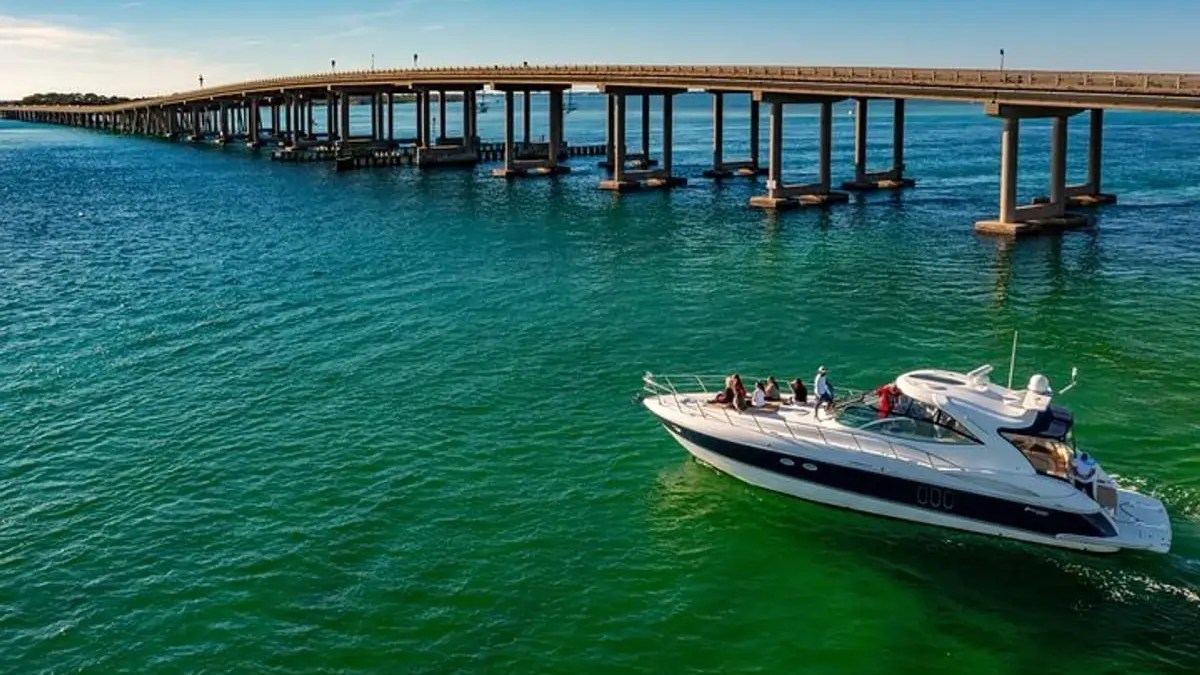 Boat with people on a calm sea under a long bridge against a clear sky.