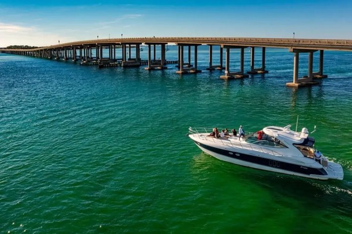 Boat with people on a calm sea under a long bridge against a clear sky.
