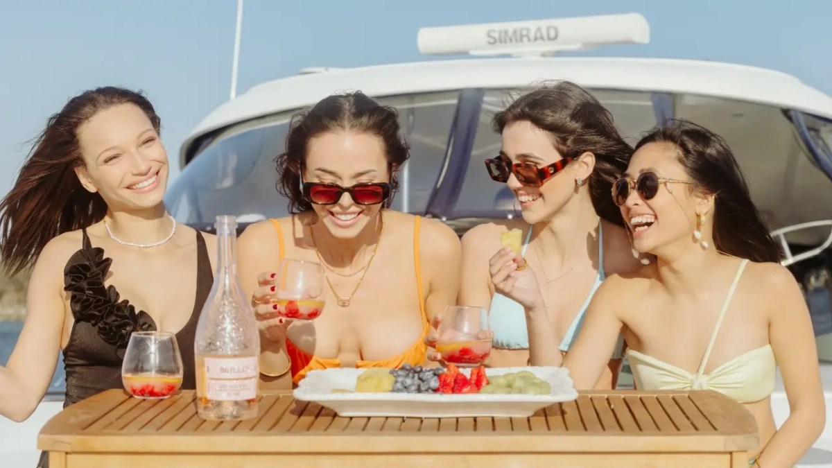 Four women in swimsuits sit on a yacht, enjoying drinks and fruit on a sunny day.