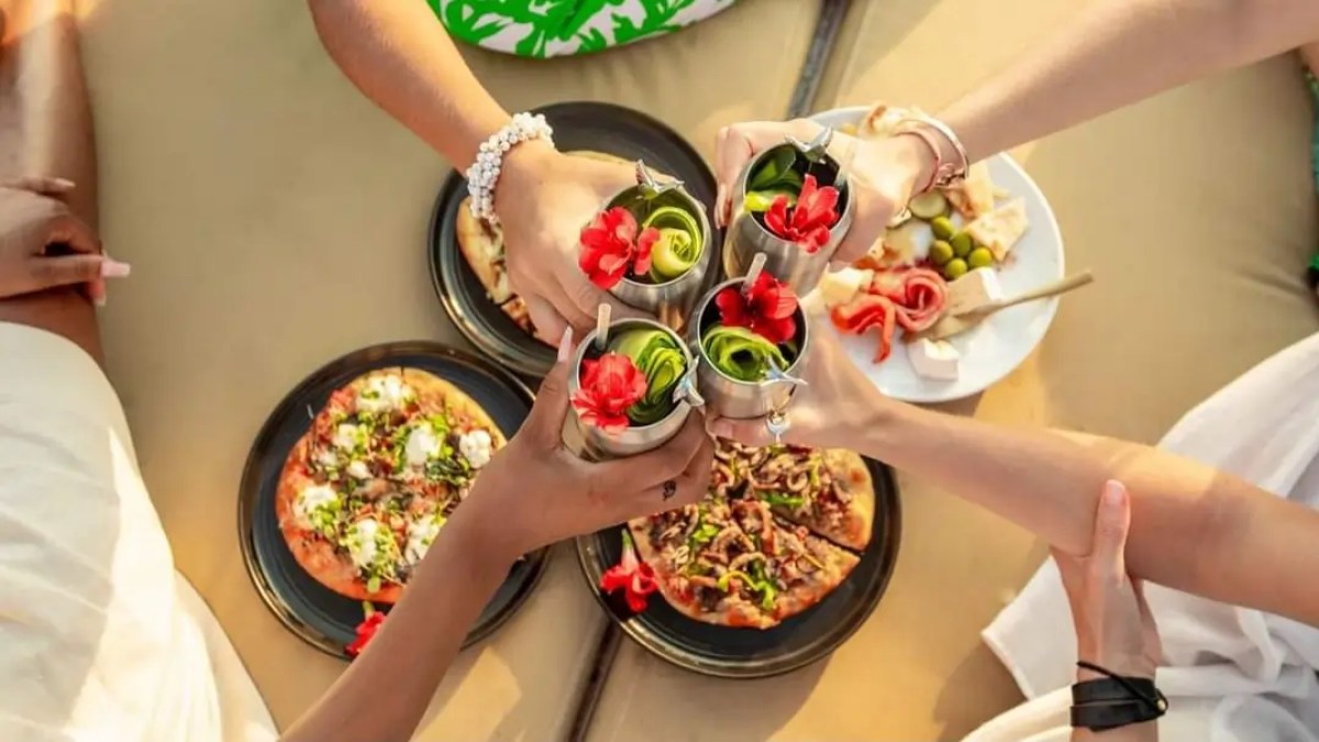 Four people toasting silver cups with cocktails above pizzas and snacks on a table.
