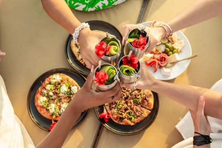 Four people toasting silver cups with cocktails above pizzas and snacks on a table.