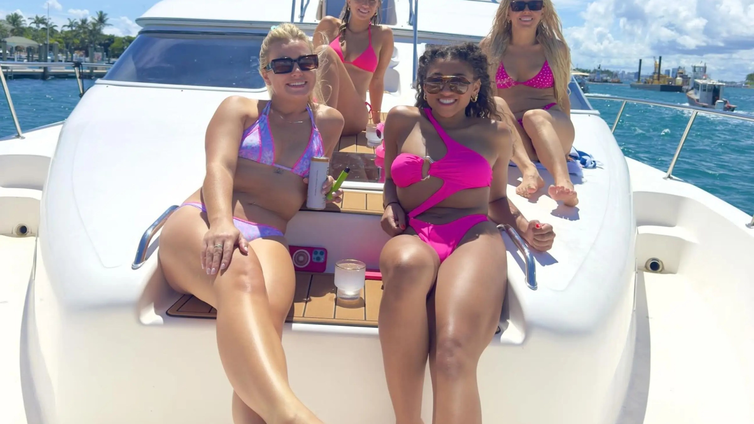 Four women in swimsuits relaxing on a yacht deck with blue sky and water in the background.