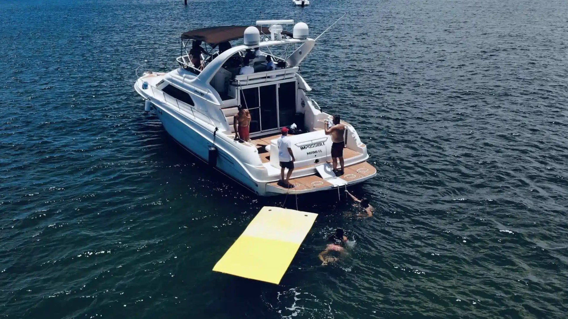 People on a yacht with a yellow floating mat, others swimming nearby in the ocean.