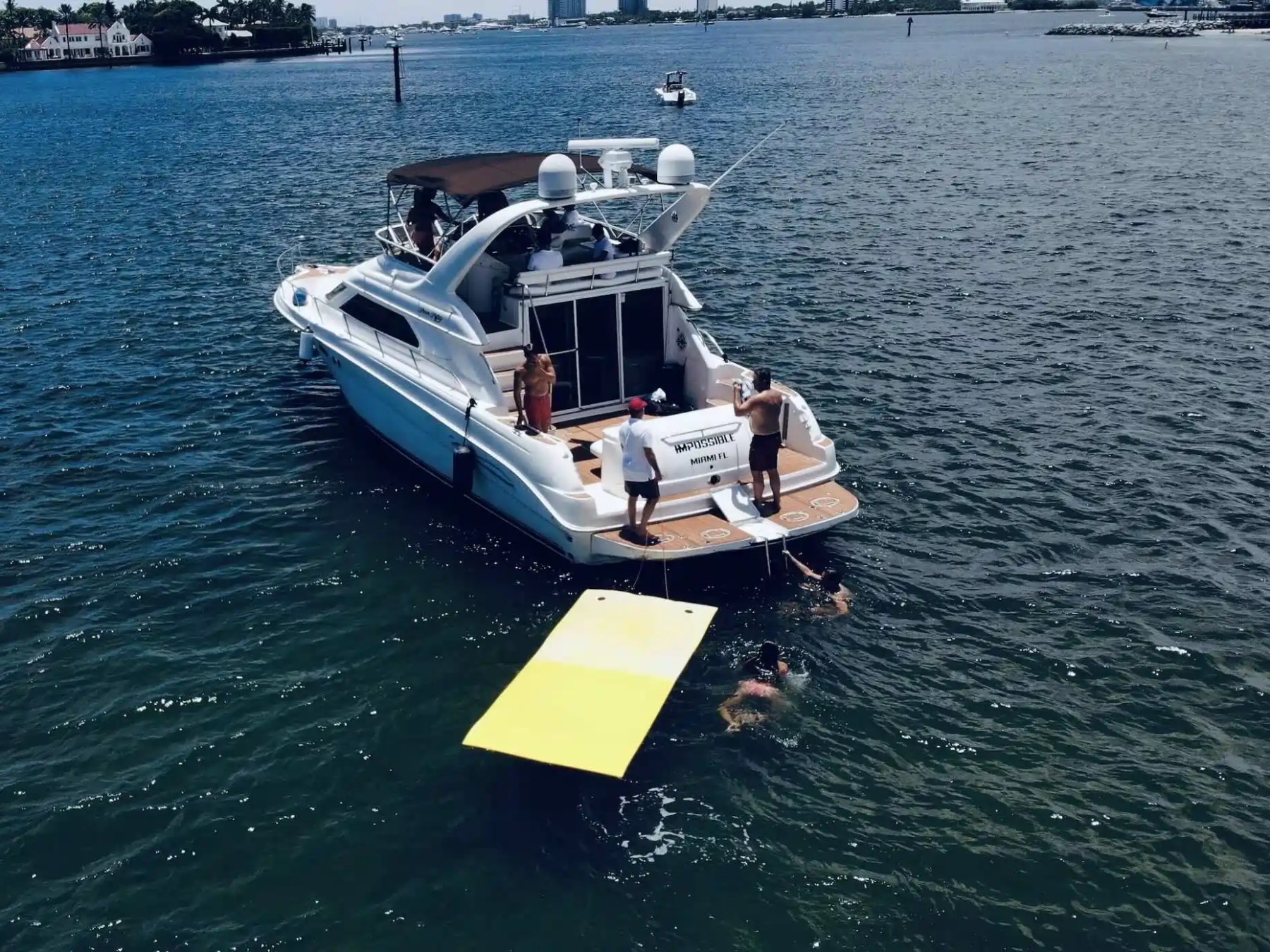 People on a yacht with a yellow floating mat, others swimming nearby in the ocean.