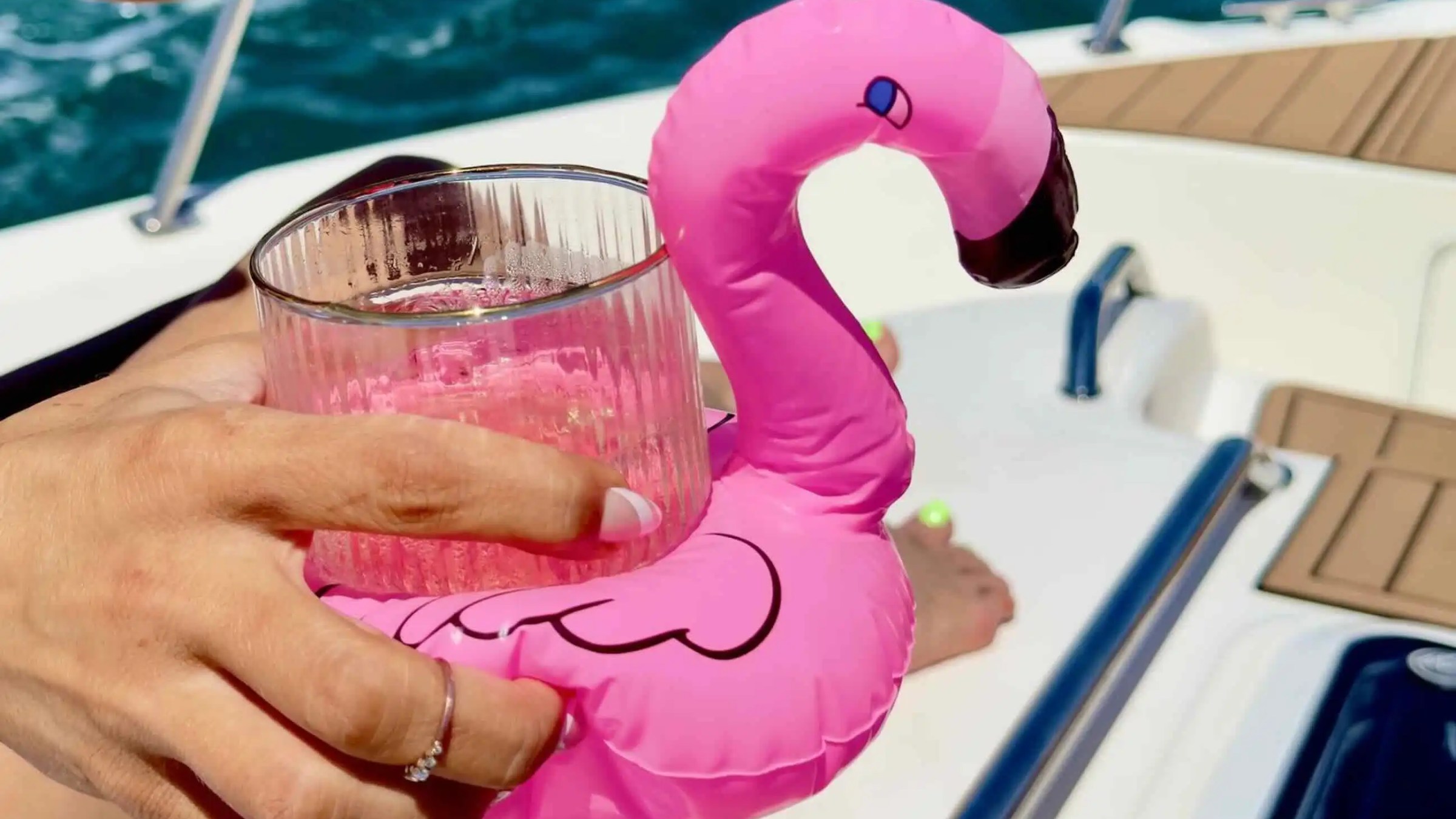 Hand holding a pink flamingo float with a glass on a boat with water background.