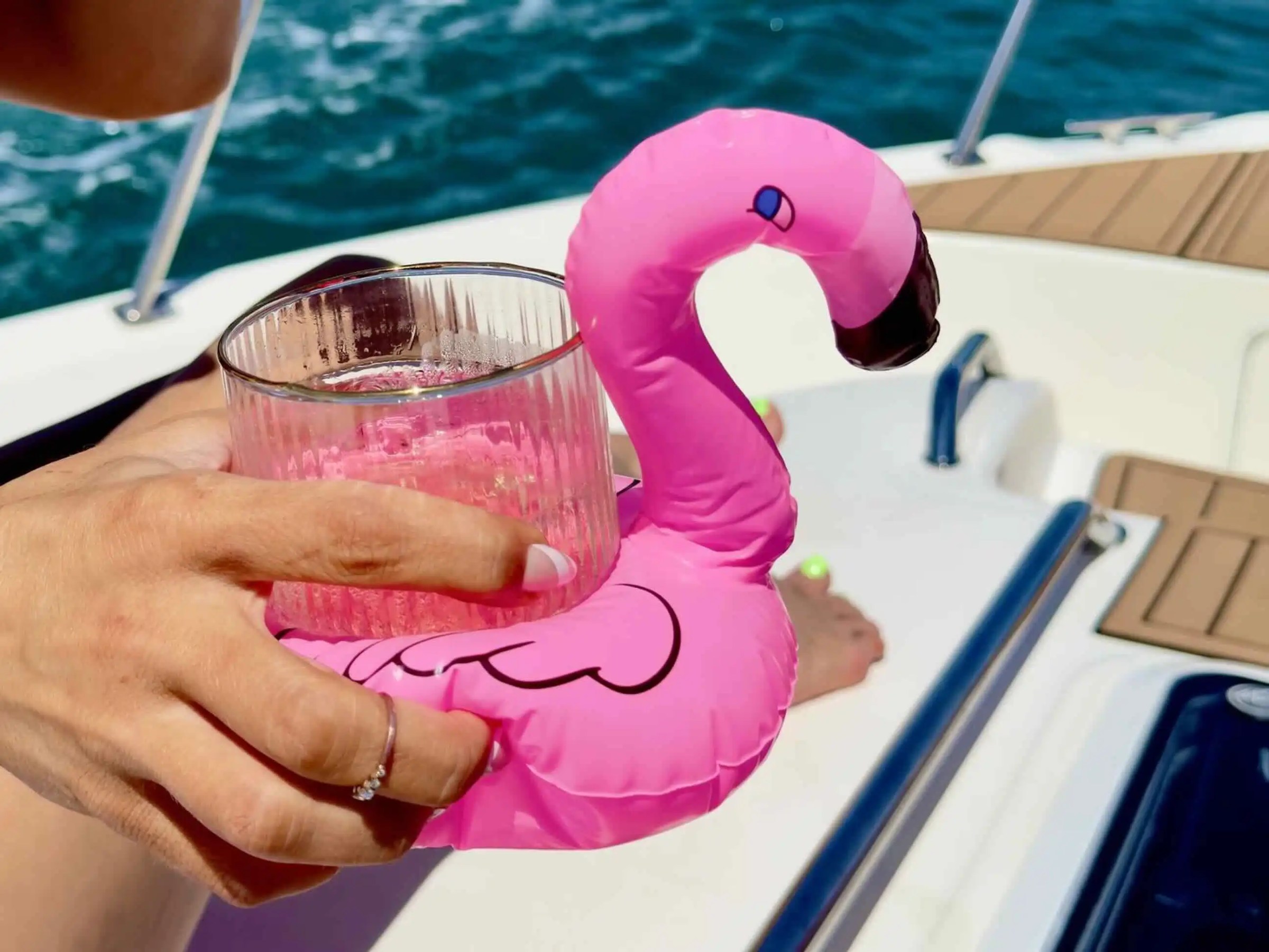 Hand holding a pink flamingo float with a glass on a boat with water background.