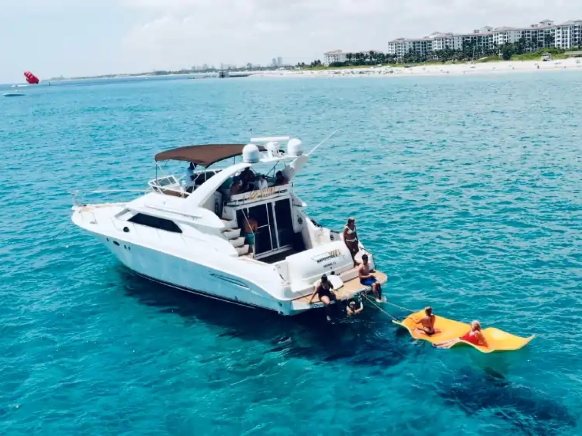 Boat with people in ocean, towing inflatable raft, near beach and buildings in background.