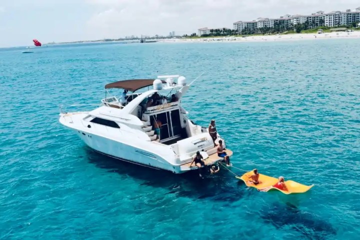 Boat with people in ocean, towing inflatable raft, near beach and buildings in background.