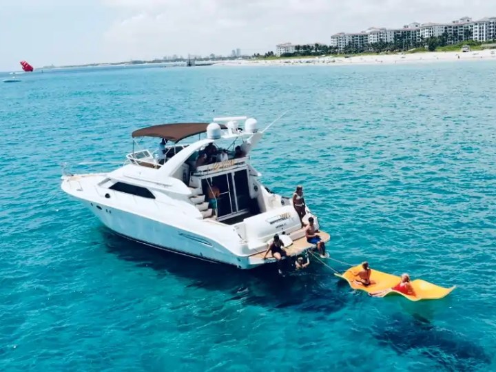 Boat with people in ocean, towing inflatable raft, near beach and buildings in background.