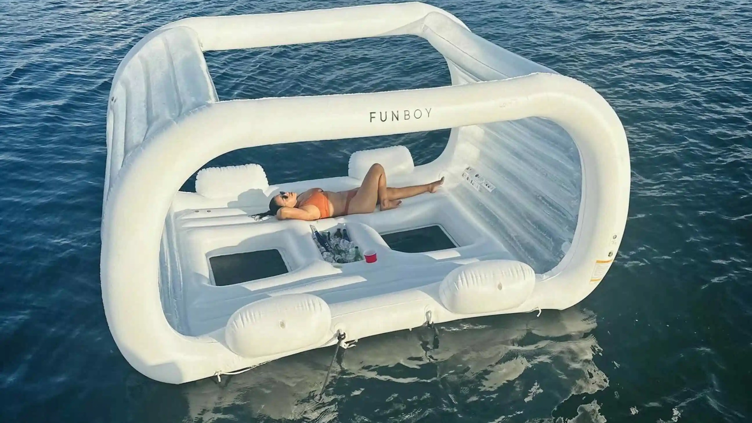 Person relaxes on a large white inflatable float with canopy on calm water.
