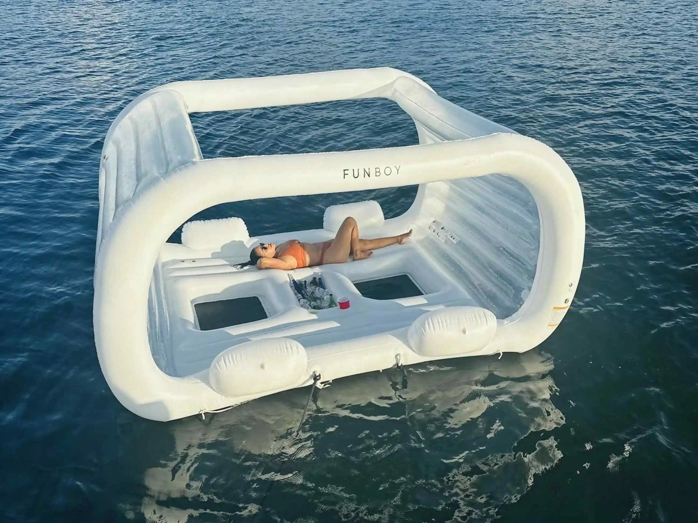 Person relaxes on a large white inflatable float with canopy on calm water.