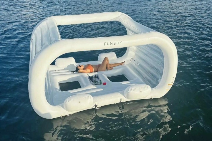 Person relaxes on a large white inflatable float with canopy on calm water.
