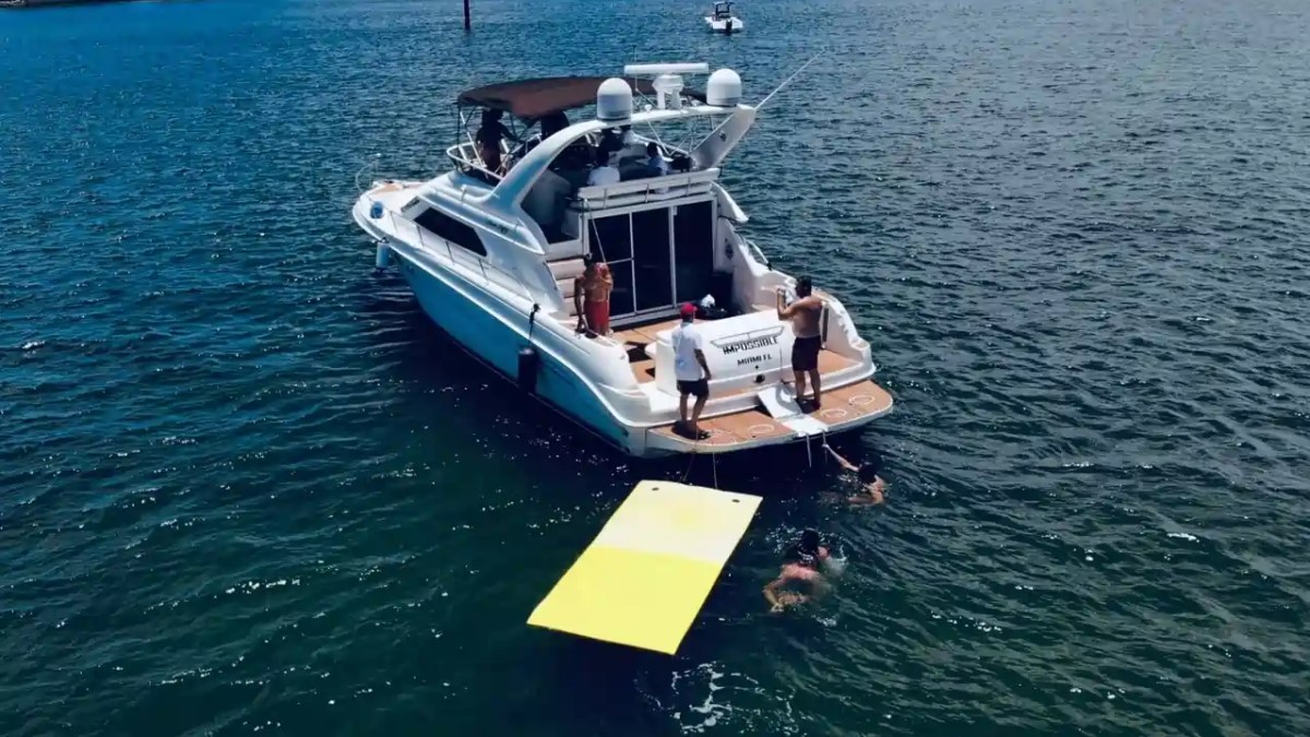 Boat in water with people swimming and a yellow floating mat nearby.