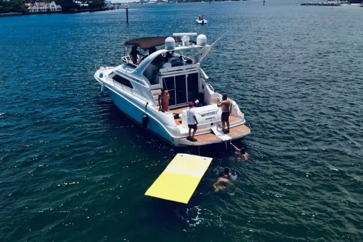 Boat in water with people swimming and a yellow floating mat nearby.