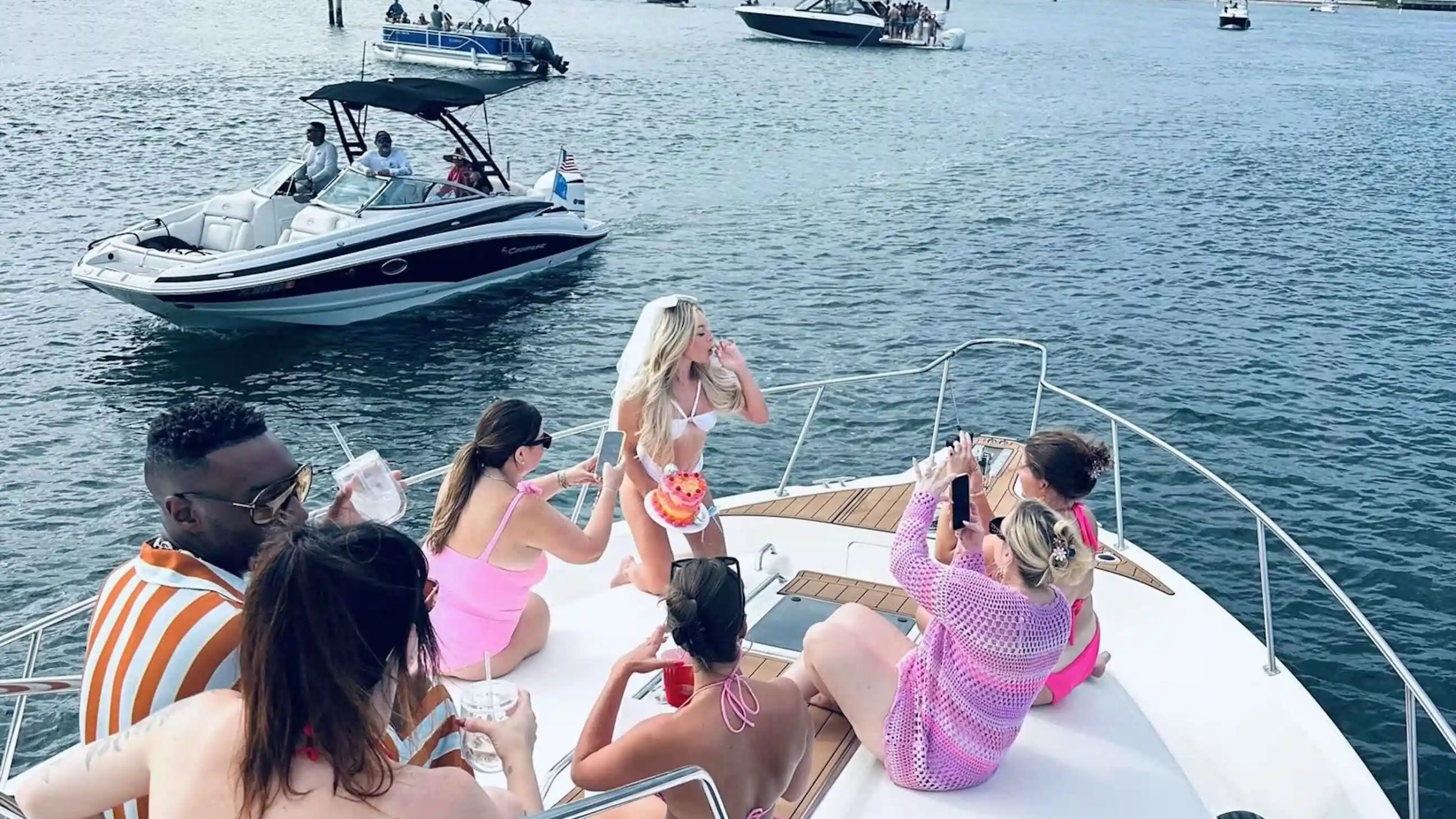 Group of people in swimwear relaxing on a boat with other boats in the background on a sunny day.