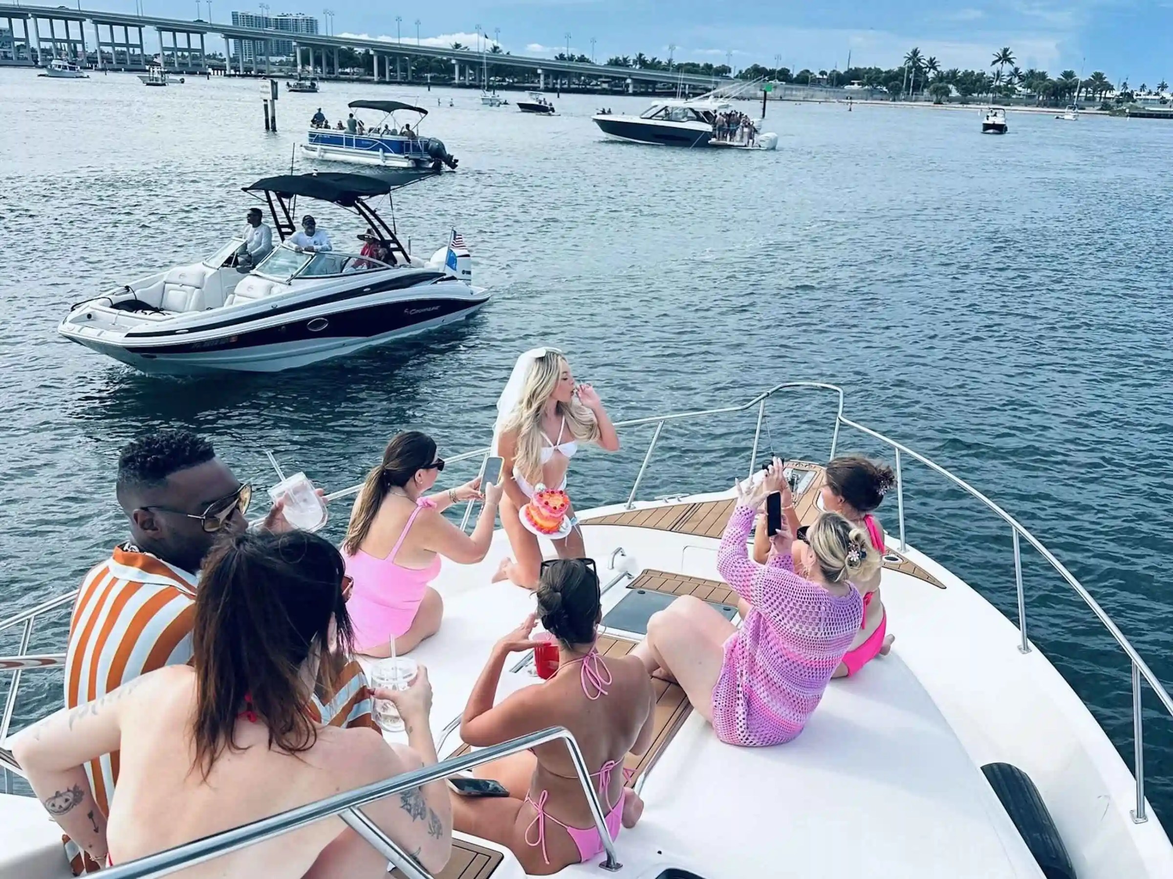 Group of people in swimwear relaxing on a boat with other boats in the background on a sunny day.